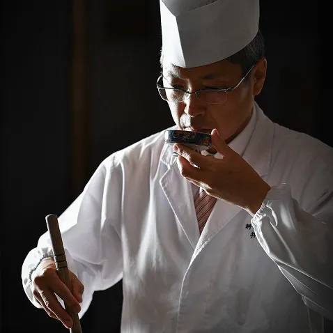 小吸物椀を手に取り、真剣な表情で出汁の香りを確かめる料理人。背景は暗く、職人の白い調理服が際立つ一枚。