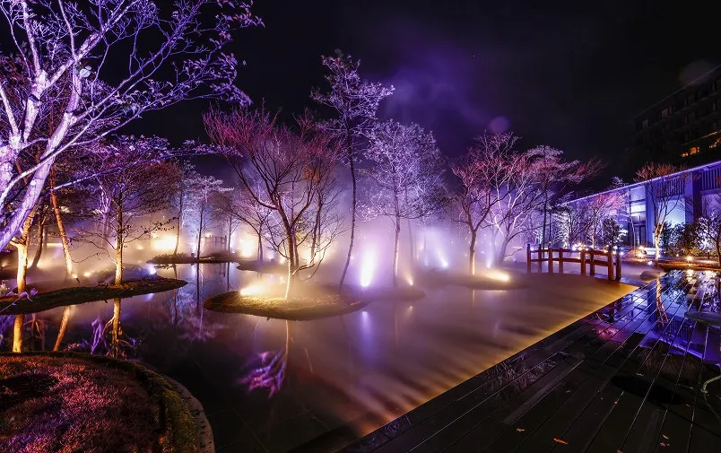 紫やオレンジの光に照らされた木々が、霧に包まれた水面に映り込む幻想的な夜の庭園。