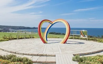 青い日本海と空を背景に立つ、カラフルな虹色のハート型オブジェ。広々とした芝生と海が一体となった開放的な風景です。
