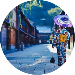 石畳が続く歴史ある古い街並みを、青い花柄の着物に黄色い帯を締めた女性が、紫色の和傘を差して一人ゆったりと歩く後ろ姿。