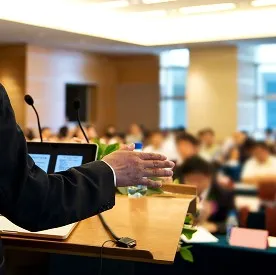 壇上でジェスチャーを交えながら講演する人物と、それを熱心に聞く多くの聴衆の後ろ姿。