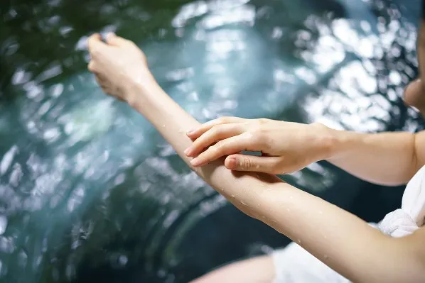 湯船の傍らで、お湯に濡れた自身の腕に優しく触れて温泉の質感を確かめる手のアップ。